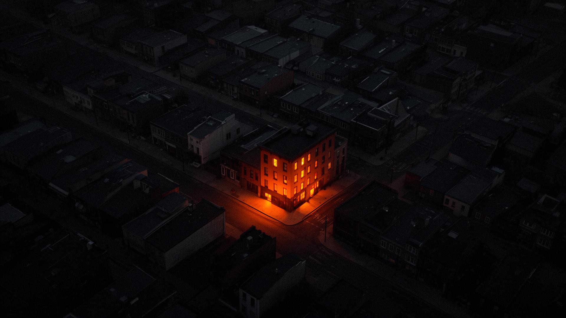 Aerial view of a dark city block at night with a single business glowing warm amber — the only one with the lights on